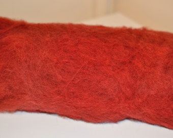 Op de afbeelding: Een close-up van een rol levendig rode wolvlies. De wol heeft een zacht, getextureerd uiterlijk, met zichtbare vezels die een pluizig, wolkachtig effect creëren. Dit materiaal is ideaal voor diverse vezelkunstprojecten, waaronder vilten en spinnen.