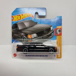 May include: A black '89 Mercedes-Benz 560 SEC AMG die-cast toy car on a blue and white Hot Wheels card. The car has white-rimmed wheels and orange headlights. The card features the Hot Wheels logo and a graphic of the car.