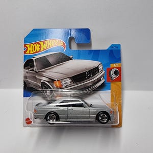 May include: A silver die-cast model car in its original packaging. The car is a coupe with a detailed design, including headlights, a grille, and wheels. The packaging features the Hot Wheels logo and the car's model name.