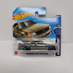 May include: A Hot Wheels die-cast toy car of a gray '89 Mercedes-Benz 560 SEC AMG. The car is in its original packaging, which is blue and features the Hot Wheels logo. The car is a 2/5 model in the series.