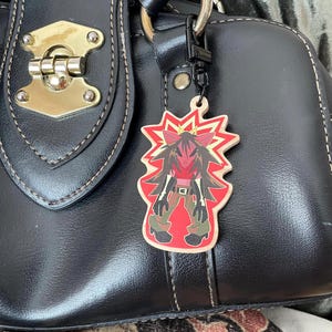 Könnte beinhalten: Ein Holzanhänger mit einer Cartoonfigur mit roten und schwarzen Haaren, einem roten Gesicht und einem roten und gelben Burst-Design. Der Anhänger ist an einer schwarzen Handtasche mit einem goldenen Verschluss befestigt.