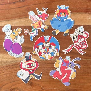 Puede incluir: Una colección de pegatinas de personajes de dibujos animados coloridos. Las pegatinas presentan varios diseños caprichosos, incluyendo un bufón, una muñeca y una criatura con alas. Cada pegatina tiene una pequeña forma rectangular azul en la parte superior.