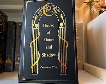 Puede incluir: Un libro de tapa dura negro con letras e ilustraciones doradas. El título es "House of Flame and Shadow" con el subtítulo "Crescent City".