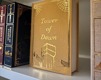 Puede incluir: Un libro de color dorado con el título "Tower of Dawn" en la portada. La portada presenta una ilustración dorada de una torre con un diseño de telaraña en las esquinas.