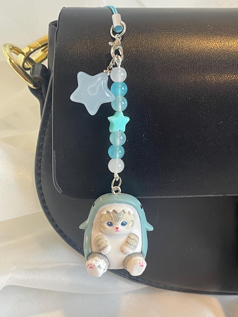 May include: A charming handbag charm with a light blue star and bead details, and a small cat figurine in a shark costume. The charm is attached to a black handbag with a gold chain handle. A delightful accessory.