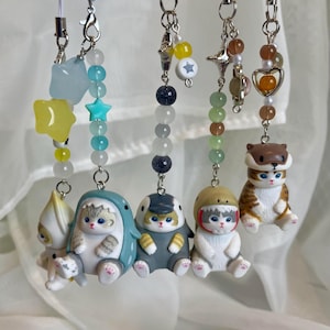 May include: Six cat-themed charms with beaded chains and silver clasps. The charms feature cats in outfits and accessories. The colours include white, blue, grey, and brown. Each charm is attached to a beaded chain.
