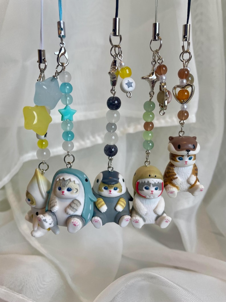 May include: Six cat-themed charms with beaded chains and silver clasps. The charms feature cats in outfits and accessories. The colours include white, blue, grey, and brown. Each charm is attached to a beaded chain.