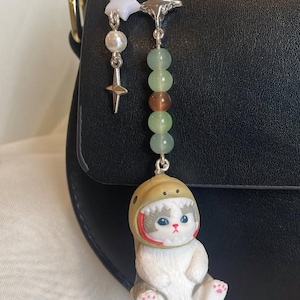 May include: A quirky cat charm wearing a space helmet, suspended from a silver chain with star and bead details. The cat figurine is white and grey with blue eyes and pink paw pads. The charm is attached to a black handbag.