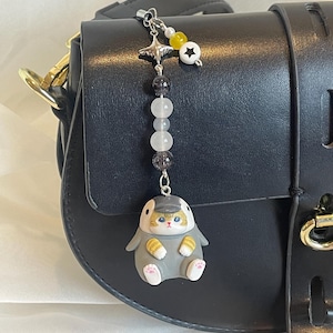 May include: A decorative charm with a grey cat-like figure wearing a hat, attached to a black handbag. The charm has a beaded chain with a star, yellow bead, and a white bead with a star. The handbag is black with gold hardware.