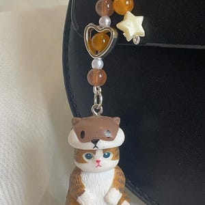 May include: A cat-themed keyring featuring a brown and white cat figurine wearing a brown hat. The keyring has a silver heart charm, star charm, and beads in brown, orange, and white. The cat has blue eyes and pink paws.