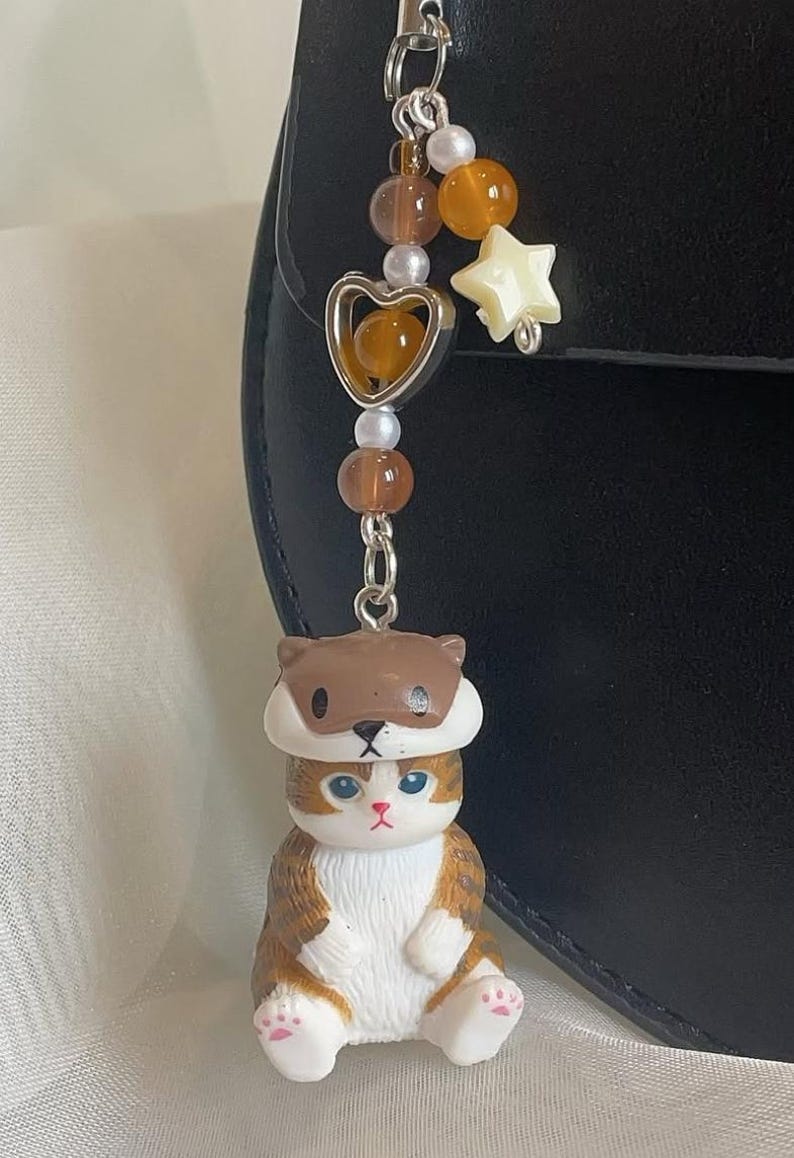 May include: A cat-themed keyring featuring a brown and white cat figurine wearing a brown hat. The keyring has a silver heart charm, star charm, and beads in brown, orange, and white. The cat has blue eyes and pink paws.