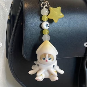 May include: A black leather handbag with a decorative charm. The charm features a cat figurine dressed as an octopus, with a yellow star and beads. The cat has a cream-coloured head and body with brown and white markings.
