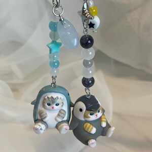 May include: Two keychains with cat figurines. One features a cat in a shark costume, the other a cat in a penguin costume. Both have silver-tone hardware and colourful beads, including star and moon charms.