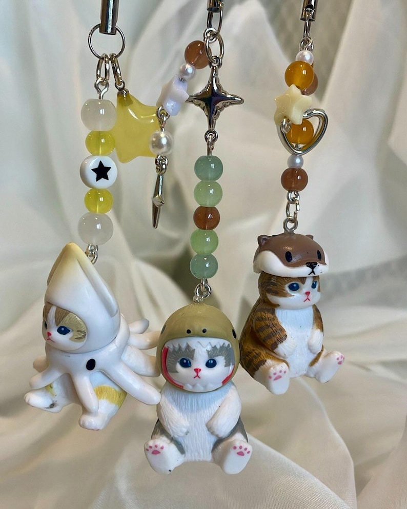 May include: Three beaded keychains with cat charms. One has a cat in an octopus costume, another a cat in a helmet, and the third a cat in a squirrel hat. Each keychain has a different colour scheme of beads and charms.
