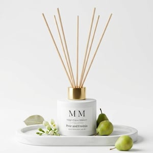 Könnte beinhalten: Ein weißer Raumduft-Diffuser mit goldfarbenem Hals und sieben Holzstäbchen. Das Etikett trägt die Aufschrift MM Magnifique Maison Pear and Freesia REED DIFFUSER | 100 ml. Er ist von zwei grünen Birnen und einem Zweig weißer Freesien umgeben.