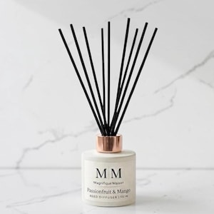 Könnte beinhalten: Ein Raumduft-Diffuser mit einer matten, weißen zylindrischen Flasche, einem metallischen roségoldenen Kragen und schwarzen Stäbchen. Das Etikett zeigt den Text MM Magnifique Maison Passionfruit & Mango REED DIFFUSER | 100 ml in schwarzer Schrift.