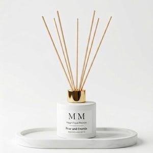 Könnte beinhalten: Ein weißer Reed-Diffusor mit goldfarbenem Kragen und Holzstäbchen. Das Etikett lautet "MM Magnifique Maison Pear and Freesia Reed Diffuser 100 ml." Der Diffusor steht auf einem weißen, ovalen Tablett.