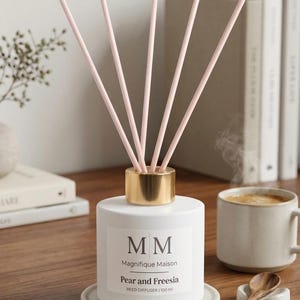 Könnte beinhalten: Weißer Schilf-Diffusor mit rosa Schilf, einem goldenen Kragen und dem Text "MM Magnifique Maison Pear and Freesia". Der Diffusor steht auf einem kleinen, runden, grauen Untersetzer. Eine Tasse Kaffee und Bücher sind im Hintergrund.