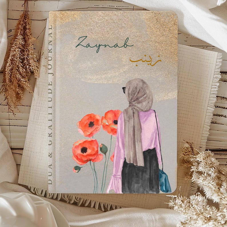 May include: A journal with the words "Dua & Gratitude Journal" printed vertically on the left side. The cover features the name "Zaynab" in English and Arabic, along with an illustration of a woman in a hijab and red poppies.