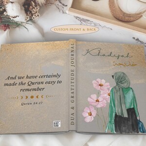 May include: Open journal with a gold and gray cover. The left side has text from the Quran. The right side features an illustration of a woman in a hijab with pink flowers. The spine reads "DUA & GRATITUDE JOURNAL."