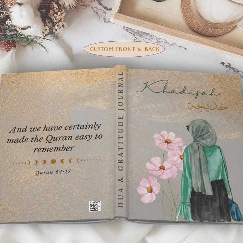 May include: Open journal with a gold and gray cover. The left side has text from the Quran. The right side features an illustration of a woman in a hijab with pink flowers. The spine reads "DUA & GRATITUDE JOURNAL."