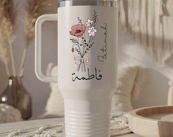 May include: A white travel mug with a handle, featuring a floral bouquet design in pink and white, tied with a bow. The name "Fatima" is written in script, along with Arabic script. The mug has a clear lid and a black top.