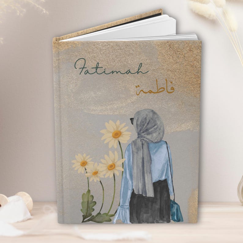 May include: A hardcover journal with the name "Fatimah" in script and Arabic calligraphy. The cover features an illustration of a woman in a hijab, daisies, and a gold and gray background.