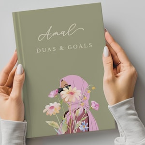 Könnte beinhalten: Ein salbeigrünes Notizbuch mit dem Titel "Amal Duas & Goals" wird gehalten. Das Cover zeigt eine Illustration einer Person mit rosa Hijab und Sonnenbrille, umgeben von Blumen. Das Notizbuch ist ein Hardcover.