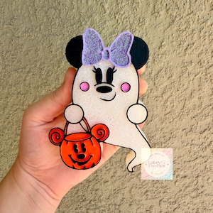 May include: A white ghost-shaped air freshener featuring black mouse ears, a glittery purple bow, pink cheeks, and a smiling face. The ghost holds an orange pumpkin-shaped bucket with a black jack-o'-lantern face. Text reads Lainey G Freshies.