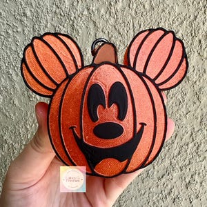 May include: A bright orange pumpkin-shaped ornament with black outlines and a black facial design resembling a cartoon character. The ornament features two smaller pumpkin-shaped ears at the top and a small brown stem. A logo reads Lainey G Freshies.