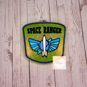 Peut inclure: Un désodorisant en forme de bouclier avec un fond vert pailleté et une bordure bleu foncé. Le texte SPACE RANGER est imprimé en noir en haut. Sous le texte se trouve un graphique d'une fusée blanche avec des ailes bleues et bleu clair.