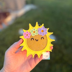 May include: A yellow sun-shaped air freshener with a textured gold center, a smiling face, and three glittery flowers in pink, green, and purple on top. A small square label with the text Lucky G Freshies is attached to the lower right side.