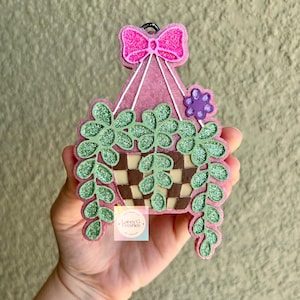 May include: A pink, glittery hanging basket-shaped air freshener with a pink bow, purple flower, and green glittery leaves. The basket has a brown and white checkered pattern. The text "Galaxy G. Freshies" is on the front.