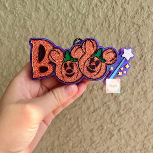 May include: A glittery orange and purple hanging ornament featuring the word BOO with two pumpkin-shaped characters resembling mouse ears, accented by a light blue magic wand with a white star and small yellow star-shaped sparkles.