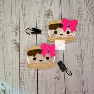 May include: Two s'mores-shaped air fresheners featuring a tan cracker, brown chocolate, and white marshmallow with a smiling face, pink cheeks, and a bright pink glittery bow. Each item includes a black metal hook for hanging and a small logo label.