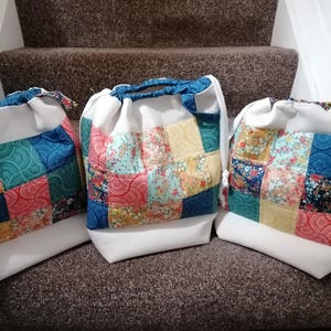 May include: Three drawstring bags made of white fabric with a patchwork design of colourful squares. The bags are lined up on a brown carpeted staircase.