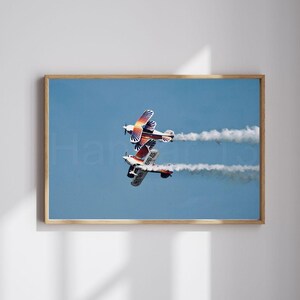 May include: A framed print featuring two biplanes in mid-air, leaving white vapor trails against a clear blue sky. The planes have colorful orange, red, and white designs. The print is in a wooden frame.