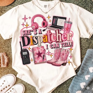 Puede incluir: Una camiseta de manga corta color crema con un estampado gráfico que incluye auriculares rosas, un portátil, un vaso, un walkie-talkie, un cuaderno y botas vaqueras rosas. El texto dice: "SHE'S A Dispatcher I CAN TELL!" con estrellas.