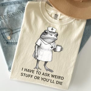 May include: A cream-colored t-shirt featuring a black and white line art illustration of a frog dressed as a nurse, holding a mug. The text below reads, I HAVE TO ASK WEIRD STUFF OR YOU'LL DIE. The shirt has a Comfort Colors brand neck label.