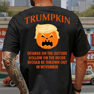 May include: A black t-shirt featuring an orange graphic of a pumpkin with a blonde hairstyle and a carved face. Above the pumpkin is the text TRUMPKIN. Below is the text ORANGE ON THE OUTSIDE HOLLOW ON THE INSIDE SHOULD BE THROWN OUT IN NOVEMBER.