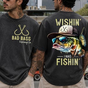 May include: A charcoal gray short-sleeve t-shirt featuring a front logo with two crossed fishing hooks and the text Bad Bass Fishing Co. The back displays a graphic of a bass wearing a baseball cap and sunglasses with the text Wishin' I Was Fishin'.