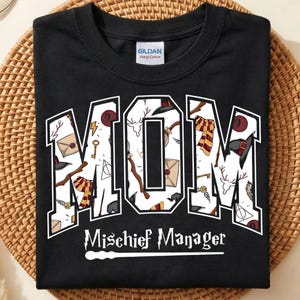 May include: A black crew neck t-shirt featuring the word MOM in large block letters filled with a pattern of wizarding-themed icons including wands, envelopes, scarves, and hats. Below, the text reads Mischief Manager with a wand graphic.
