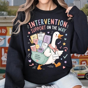 May include: A black crewneck sweatshirt featuring a graphic of a white duck carrying a teal bag filled with school supplies, including books labeled RTI, Reading Cards, Math Cards, and Data Tracker. The text reads Intervention Support On The Way.