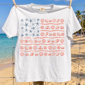 May include: A white short-sleeve t-shirt featuring a graphic print of an American flag composed of blue starfish and sand dollars in the upper left corner, with horizontal stripes made of various orange seashells across the rest of the shirt.