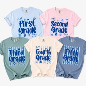 以下が含まれることがあります： ライトブルー、ピンク、セージグリーン、クリーム、スレートブルーの半袖Tシャツ5枚。各シャツには、縦縞、星、花、リボンの青いグラフィックが描かれており、first grade、second grade、third grade、fourth grade、fifth gradeという文字がプリントされています。