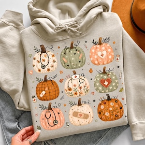 Op de afbeelding: Een beige hoodie met een grafische print van negen pompoenen versierd met medische thema's, waaronder stethoscopen, verpleegsterskapjes, pleisters en bloemmotieven, afgewisseld met kleine iconen van medicijnflesjes en thermometers.