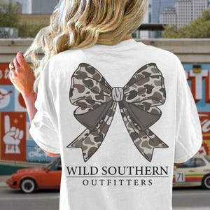 Könnte beinhalten: Ein weißes T-Shirt mit einer großen Grafik auf der Rückseite, die eine Schleife mit braun-grauem Tarnmuster zeigt. Unter der Schleife steht der Text WILD SOUTHERN OUTFITTERS in schwarzen Großbuchstaben, eingerahmt von einer Linie darüber und darunter.
