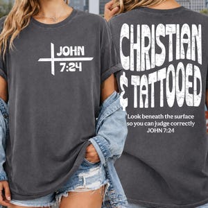 May include: A charcoal gray t-shirt featuring white text. The front displays a cross graphic with the text JOHN 7:24. The back features the text CHRISTIAN & TATTOOED in a large distressed font, followed by the text Look beneath the surface so you can judge correctly JOHN 7:24.