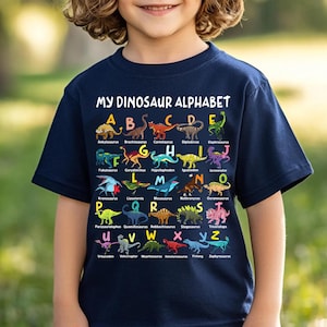 May include: A navy blue short-sleeved t-shirt featuring a colorful graphic print titled MY DINOSAUR ALPHABET. The design displays letters A through Z, each paired with a unique dinosaur illustration and its corresponding name printed in white text.