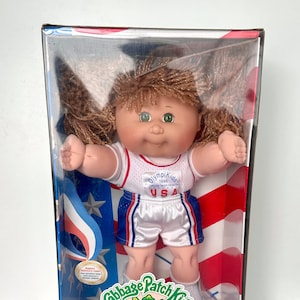 May include: A Cabbage Patch Kids OlympiKids Special Edition doll in its original box. The doll has brown hair, green eyes, and is wearing a white and red USA team uniform. The box features patriotic colors and the OlympiKids logo.